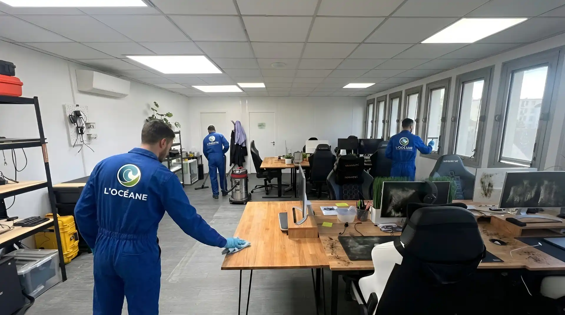 3 agents de nettoyage en uniforme bleu avec le logo L'OCÉANE dans le dos sont en train de nettoyer un local professionnel pour un tarif. Ils essuient les bureaux, nettoient le sol et lavent les vitres. Le bureau est doté de grandes fenêtres, de bureaux, de chaises et d'ordinateurs.